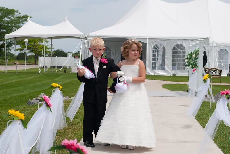 Two children walk together along a paved aisle edged with white fabric and pink and yellow flowers.