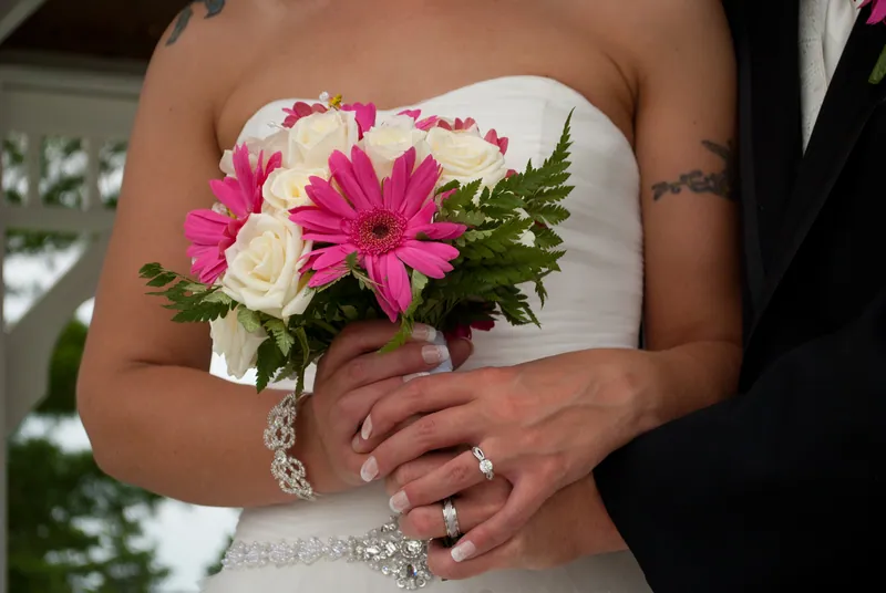 Angela holds a bouquet of bright pink daisy-shaped flowers, ivory roses and green foliage against her white dress.