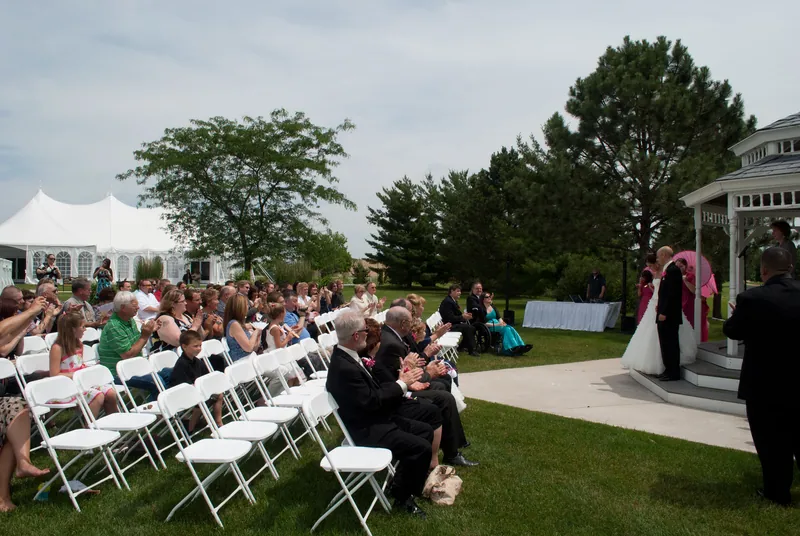 Rows of seated guests face the gazebo across a lawn, with an open paved area at the front.