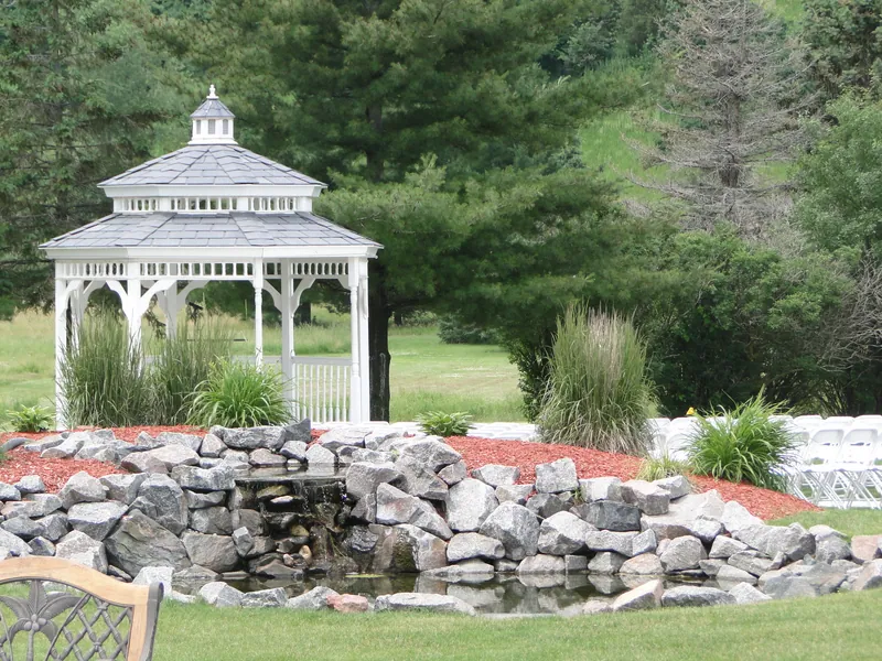 A white gazebo stands beyond a stone water feature, surrounded by grass and mature trees.