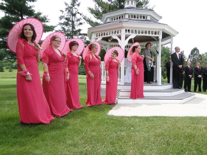 Wedding-party members in vivid pink dresses hold matching pink parasols beside the white gazebo.