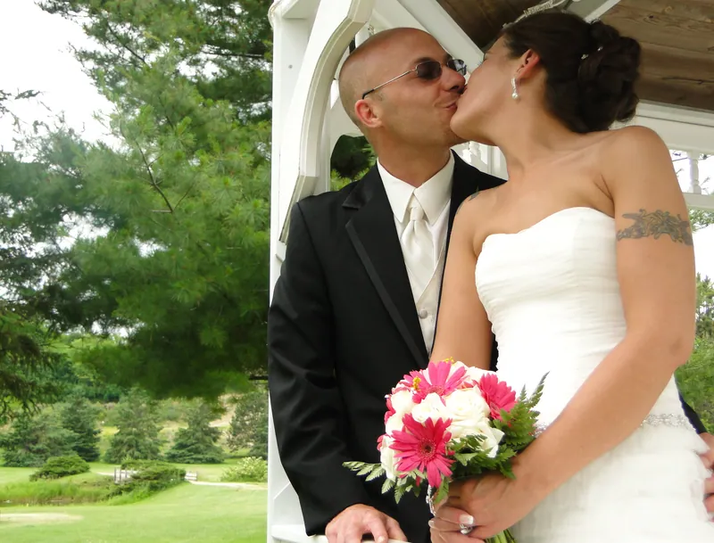 Angela and Jeremiah kiss beside a white gazebo, with a bright pink and ivory bouquet in the foreground.