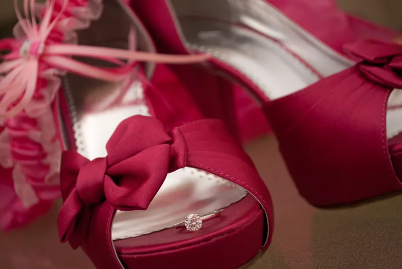 Deep pink open-toe shoes with fabric bows sit beside pale pink ribbon, with a ring in the foreground.