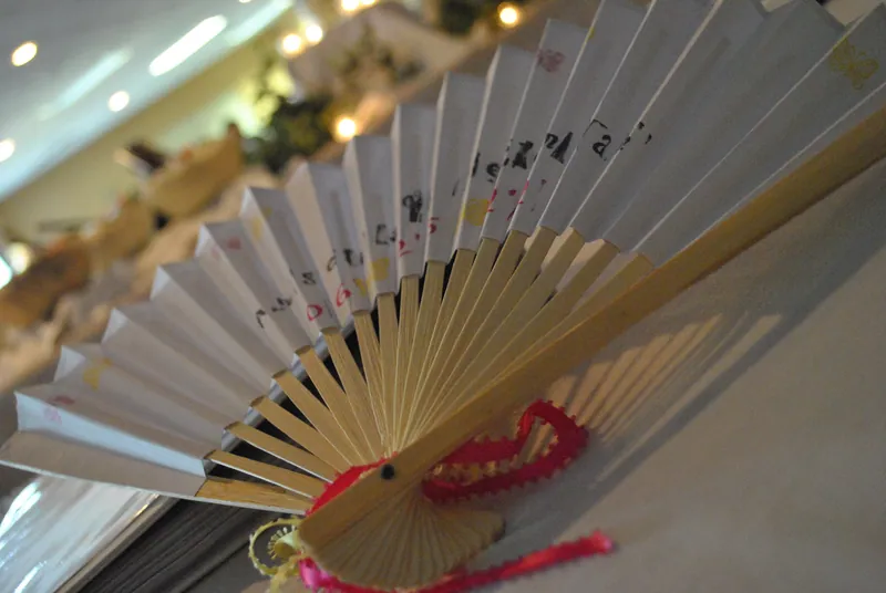 An open folding fan with pale panels and a pink cord rests on a reception table.