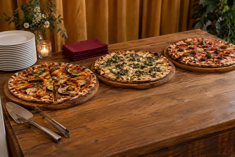Three boards of sliced vegetable pizza, clean serving utensils and a plate stack form a compact indoor evening-food station.