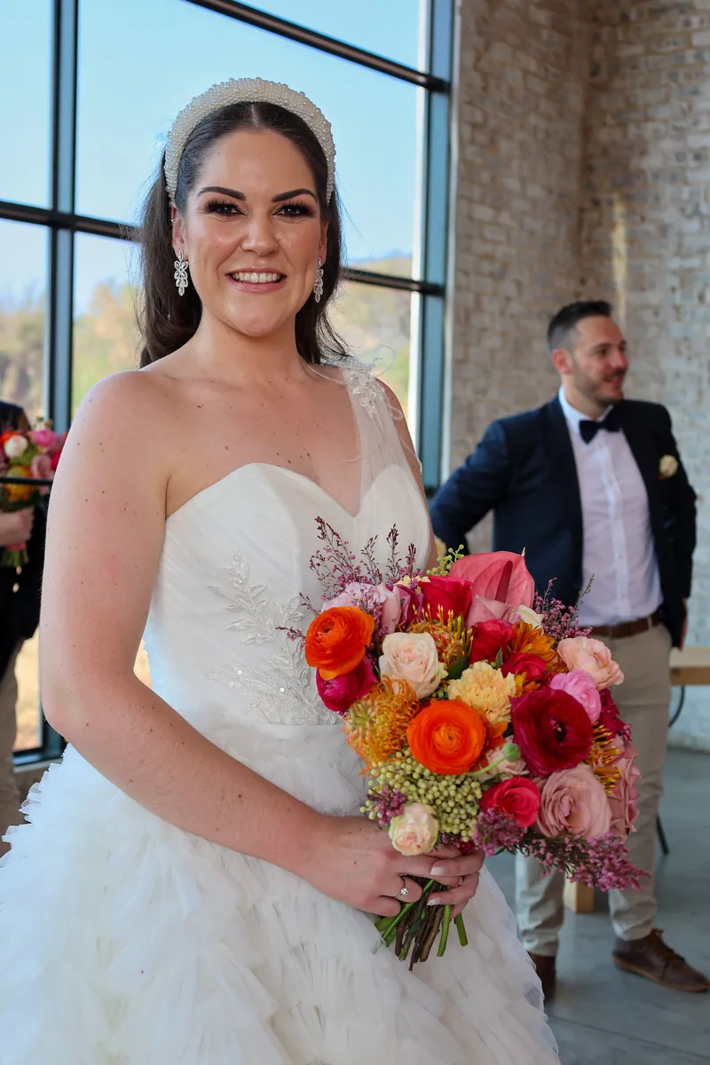 Roxy holds a rounded bouquet of pink, orange, yellow and pale flowers against her ivory gown.