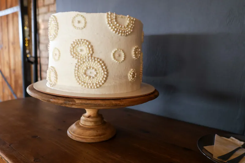 An ivory single-tier cake with pearl-like circular decorations stands on a wooden pedestal beside a dark wall.