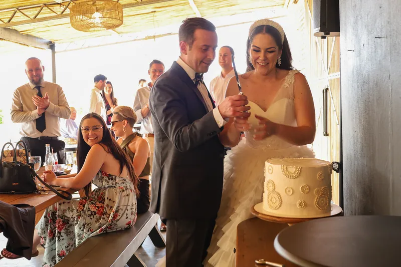 Roxy and Matthew stand together at the cake, holding a knife beside the ivory cake.
