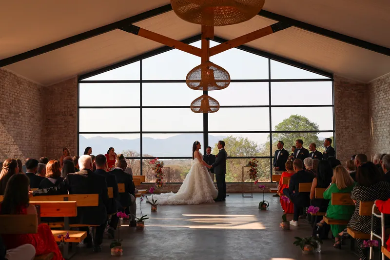 Guests sit on timber benches in a brick ceremony room with a large gridded window behind the couple.