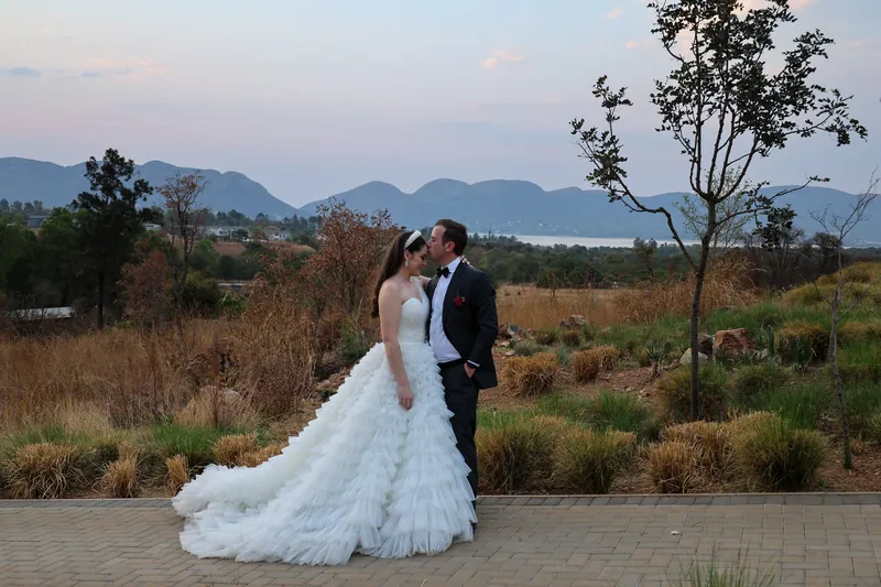 The couple stand together on a paved path beside grasses, with hills and water in the distance.